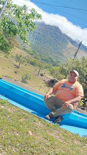 A man sitting on a blue slide in a grassy area with a mountainous background.