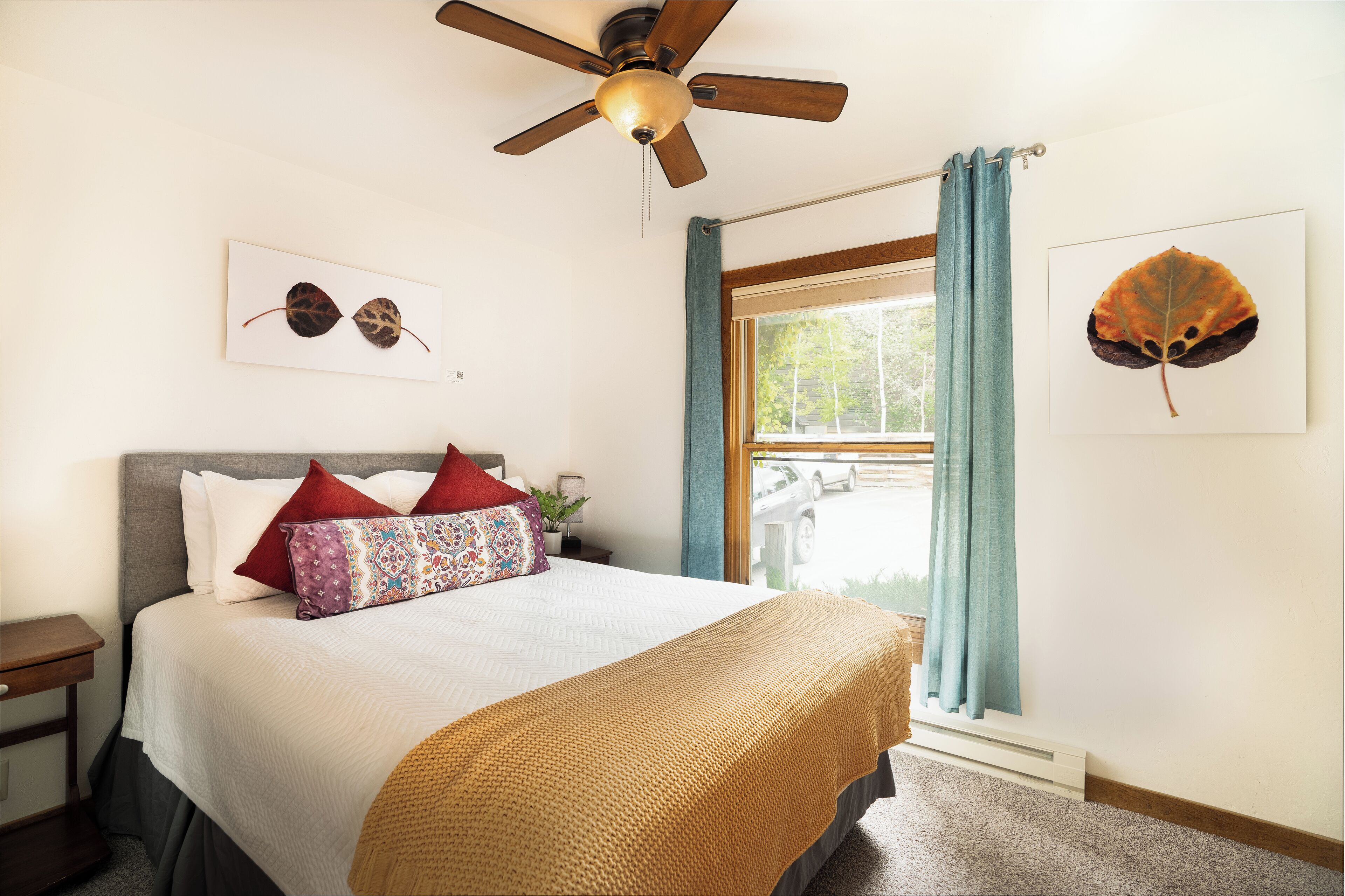 A neatly made bed with a patterned pillow, a ceiling fan, and two framed leaf artworks on the walls.