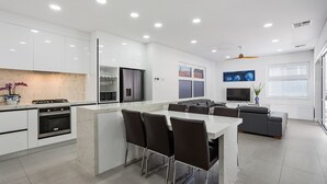 A modern kitchen with white cabinets, a central island with bar stools, a built-in oven, and a dining area with a gray sofa and TV.
