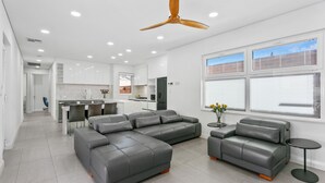 A modern living room with grey leather sofas, a ceiling fan, and large windows with blinds.