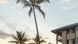 A poolside view with lounge chairs, palm trees, and a building with balconies overlooking the ocean at sunset.