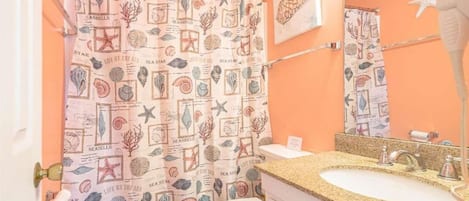 A bathroom with a seashore-themed shower curtain, a white toilet, a granite countertop sink, and a hairdryer on the wall.