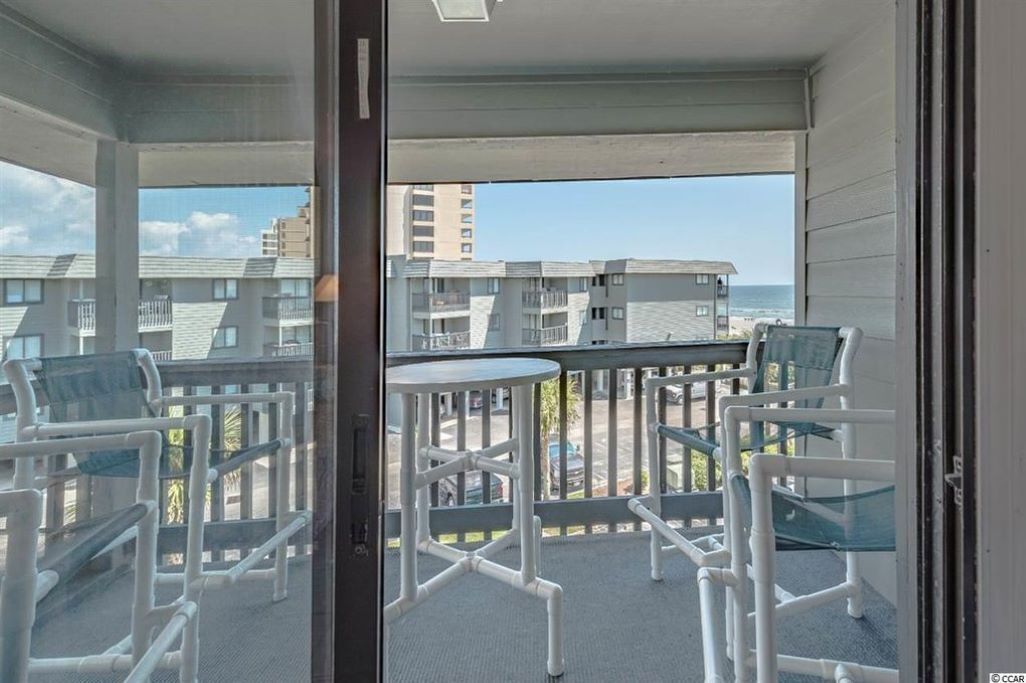 A balcony with white chairs and a small table, overlooking a beach and other buildings.