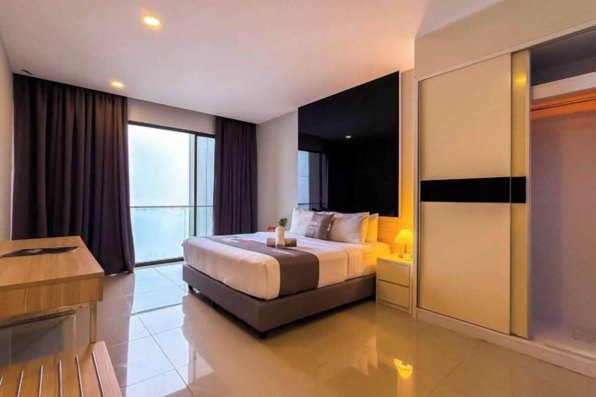 A modern hotel room with a large bed, a desk, a nightstand, and a sliding door closet.