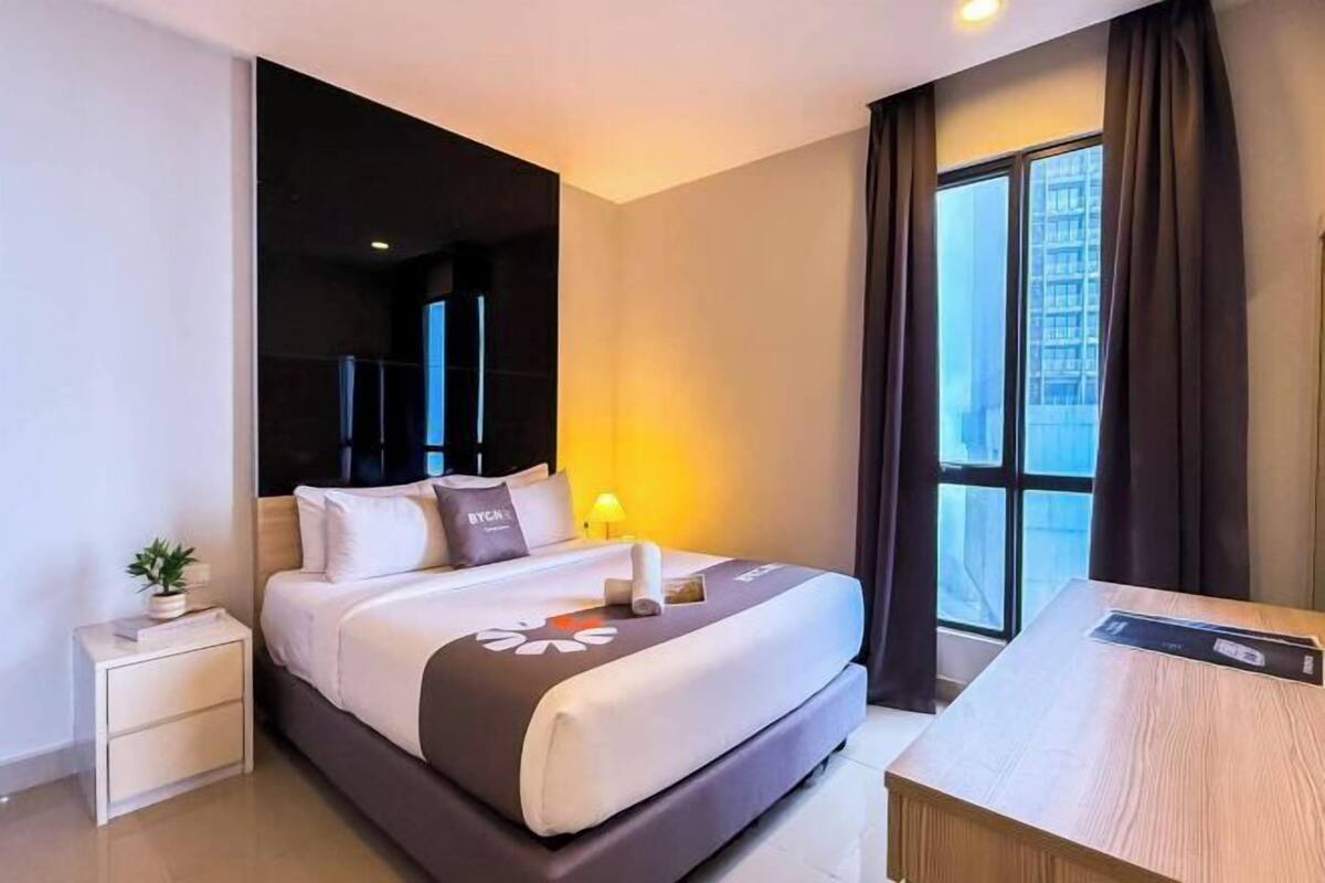 A hotel room with a bed, bedside table, lamp, and a view of the city through large windows.
