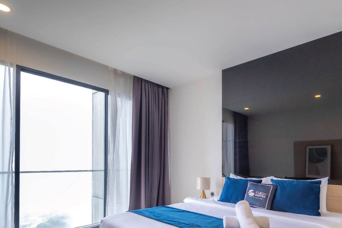 A hotel room with a large bed, a window with curtains, a mirror, and a wall-mounted light.