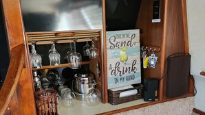 A wooden bar area with multiple clocks, a sign reading "Toes in the Sand - A drink in my hand," and various glassware and bottles.