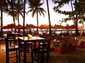 Un área de comedor al aire libre con mesas y sillas, rodeada de palmeras y vistas a la playa.
