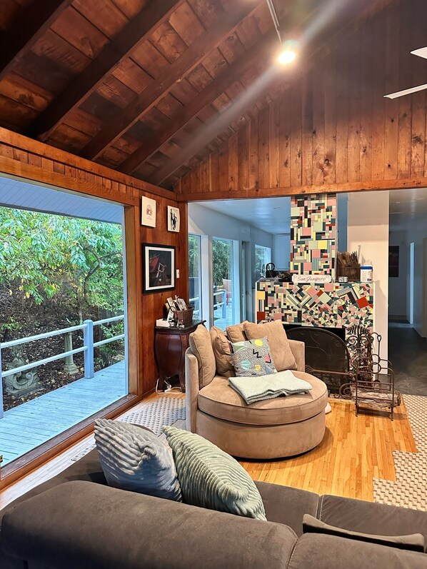 Un salon chaleureux avec un plafond en bois, un canapé rond, une cheminée et de grandes fenêtres donnant sur un jardin.