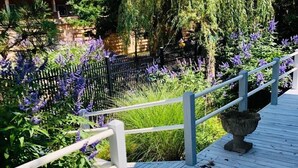 Una terraza de madera con baranda blanca, rodeada de exuberante follaje y flores moradas.