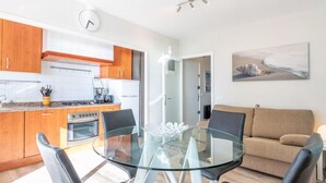 Eine moderne Küche mit Holzschränken, einem Gasherd und einem Kühlschrank. Es gibt einen Essbereich mit einem Glastisch und vier Stühlen. Im Wohnzimmer steht ein beiges Sofa, und ein Gemälde hängt an der Wand.