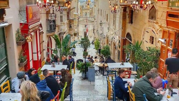 Une rue étroite bordée de cafés avec des tables en terrasse, de bâtiments en pierre et d’ampoules suspendues.