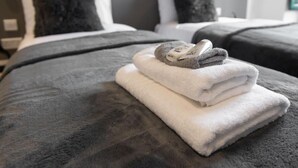 Una cama bien hecha con una manta gris y una toalla blanca doblada, con un par de pantuflas encima.