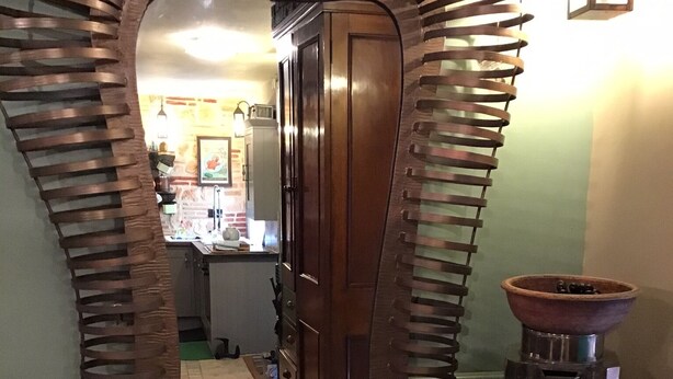 A mirror with a unique ribbed frame, a wooden door, and a kitchen area with a coffee machine and a bowl on a small table.