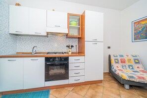 A compact kitchen with white cabinets, a stove, and a microwave. There's a small shelf with dishes and a colorful artwork on the wall.