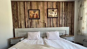 Cama doble con sábanas blancas, dos almohadas y una manta beige con textura. Hay dos mesitas de noche con lámparas, y paneles de madera en la pared detrás de la cama.