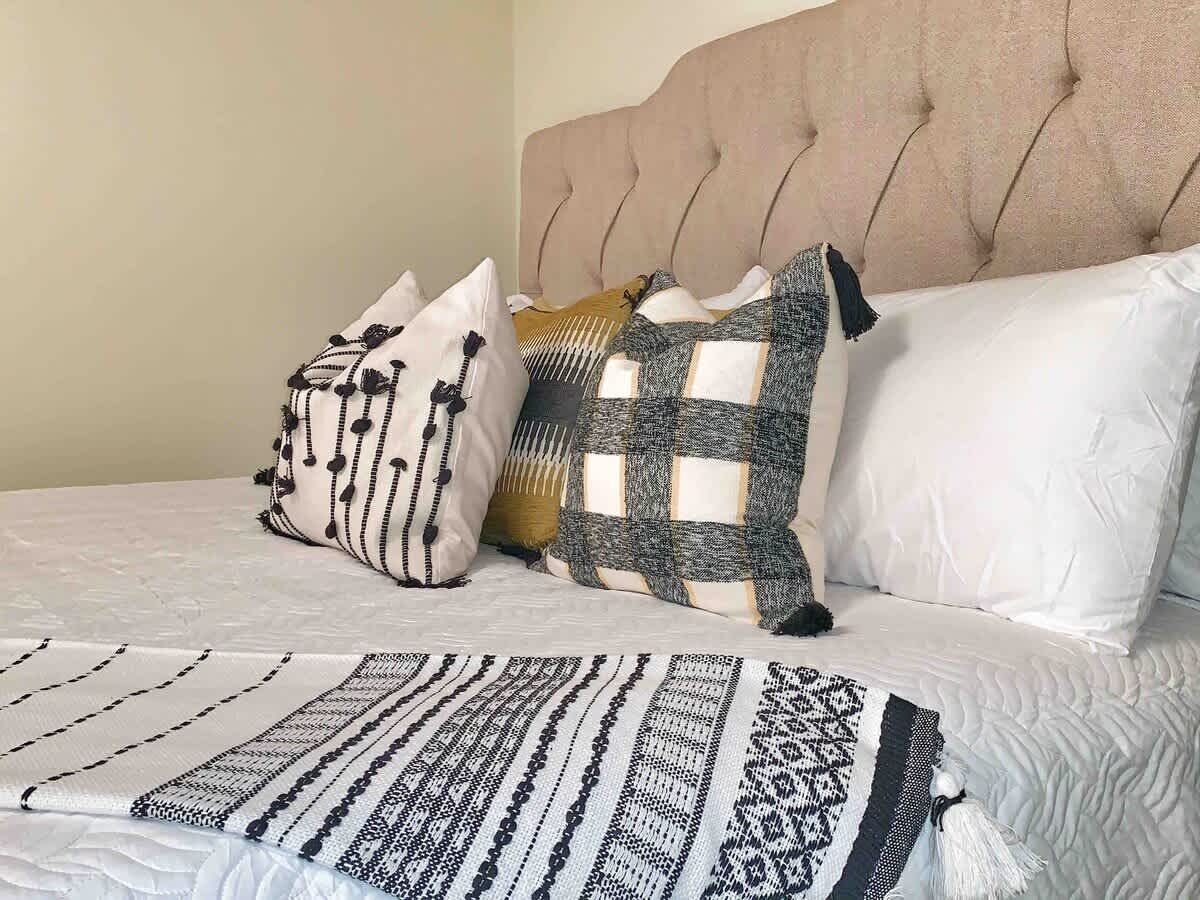 A bed with a tufted headboard, a patterned throw blanket, and three pillows with different designs.