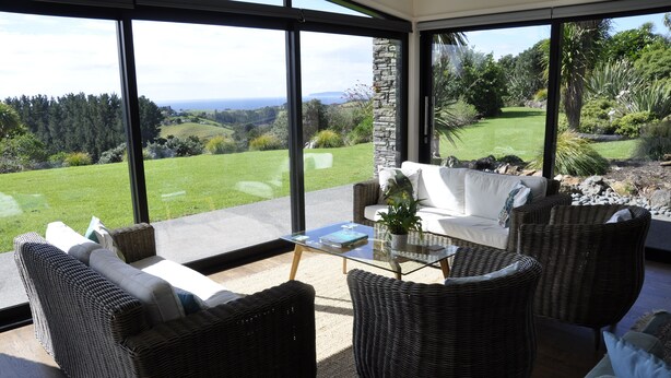 A modern living room with wicker furniture, a glass coffee table, and a view of a lush garden and distant landscape.