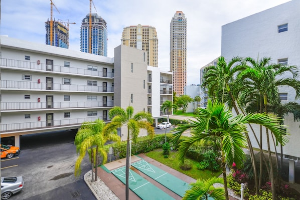 A multi-story residential building with balconies, a parking area, and a green space with palm trees.