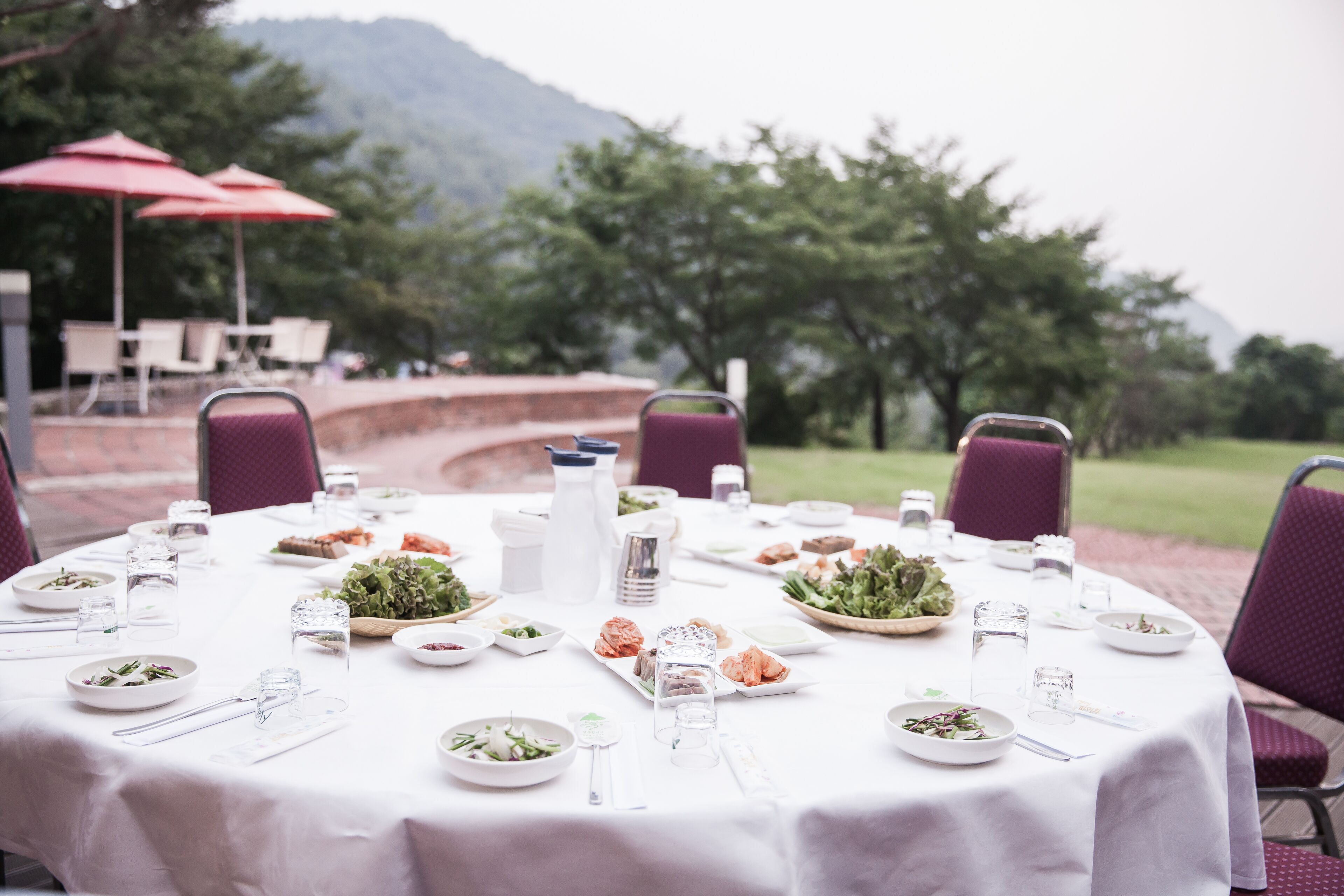 An outdoor dining table set with plates, glasses, and cutlery, surrounded by chairs and red umbrellas, with a view of trees and a mountain in the background.