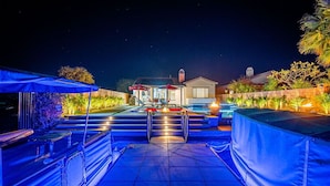 Vista nocturna de un patio trasero residencial con piscina, escalones que llevan a una casa y zonas de estar al aire libre.