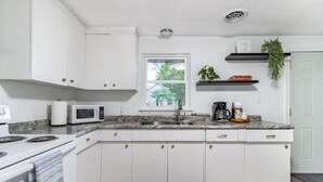 Una cocina moderna con armarios blancos, encimera de granito, microondas integrado, estufa, lavamanos y ventana con vistas al exterior.