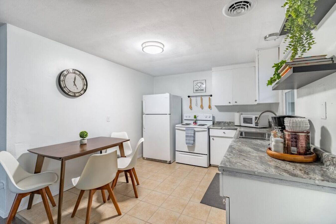 Una cocina compacta con armarios blancos, estufa, microondas y lavamanos. Hay un reloj en la pared y una pequeña planta en la encimera.
