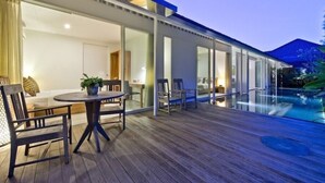 A modern outdoor deck with a pool, wooden furniture, and glass doors leading to an interior space.