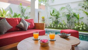 A poolside setting with a red sofa, two glasses of orange juice, and a bowl of fruit on a wooden table. - Villa Double 6 (Seminyak)
