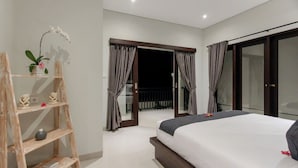 A bedroom with a bed, a wooden shelf with decorative items, a potted plant, and a sliding glass door leading to an outdoor area. - Villa Double 6 (Seminyak)