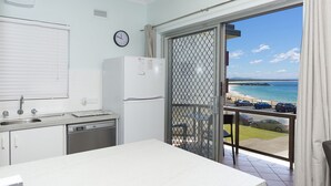 A kitchen with a white countertop, a sink, and a refrigerator. There is a window with a view of the beach and ocean. - Aloha Beachside (Forster)