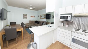 A compact kitchen with white cabinets, a microwave, and a stove. There is a dining area with a table and chairs, and a sofa in the background. - Aloha Beachside (Forster)