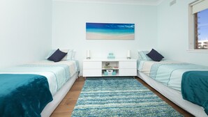 A bedroom with two beds, a nightstand, a painting of a beach, and a view of the city through a window. - Aloha Beachside (Forster)