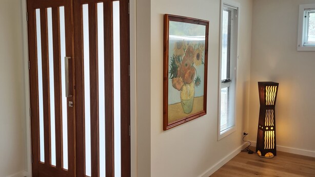 Ein Zimmer mit Holzboden, einer großen Holztür mit vertikalen Lamellen, einem gerahmten Gemälde an der Wand und einer Stehlampe.