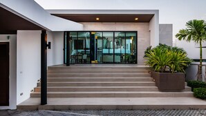Um edifício moderno com escadaria que leva a uma porta de vidro, um jardim elevado de concreto com plantas e uma área pavimentada com grade metálica.