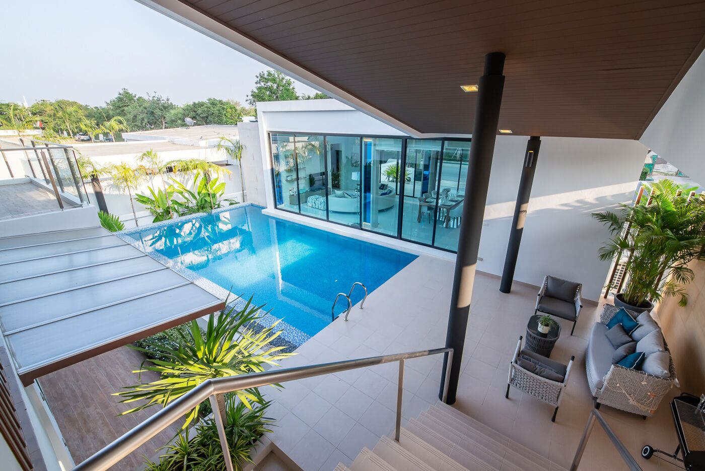 A modern outdoor pool area with a glass enclosure, a staircase leading to a deck, and a seating area with a sofa and chairs.
