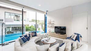 Uma sala de estar moderna com um sofá branco, uma mesa de centro e uma TV de tela plana. Há uma janela grande com vista para uma piscina e uma varanda.