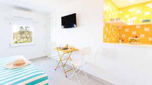 Una habitación luminosa con una cama, una mesa con sillas, un televisor y una zona de cocina con salpicadero de azulejos.