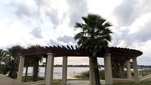 Une structure de pergola en bois, avec des palmiers au premier plan, surplombant une étendue d’eau dont on aperçoit la rive au loin.