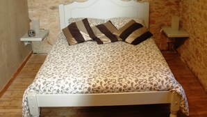 A bed with a patterned bedspread and two pillows, a white headboard, and a stone wall background. - Time for a break - the barn (Berbiguières)