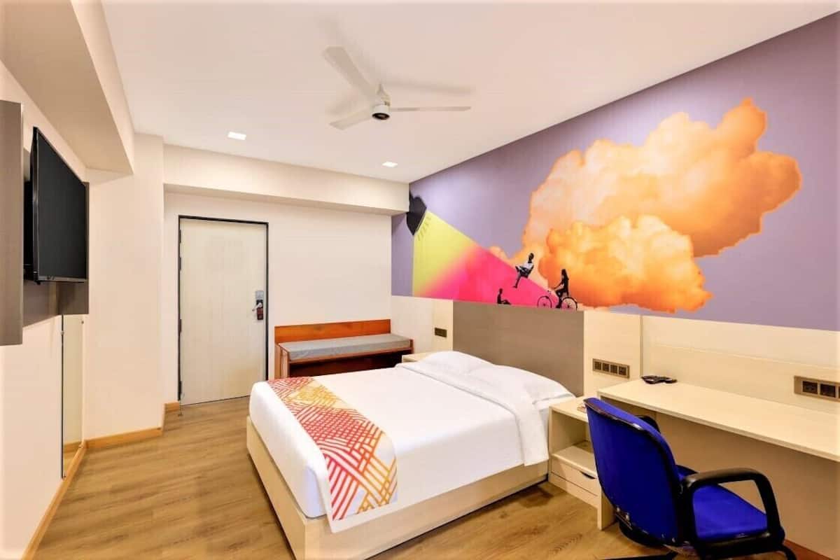 Quarto de hotel com cama, escrivaninha, cadeira, ventoinha no teto e um mural na parede.