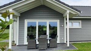 Een huis met een grijze gevel, witte raamkozijnen, een overdekte veranda met rieten meubels, en een grote schuifpui.