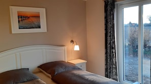 Une chambre à coucher avec un lit, des oreillers, une table de chevet, une lampe fixée au mur, un tableau encadré et une fenêtre avec des rideaux.