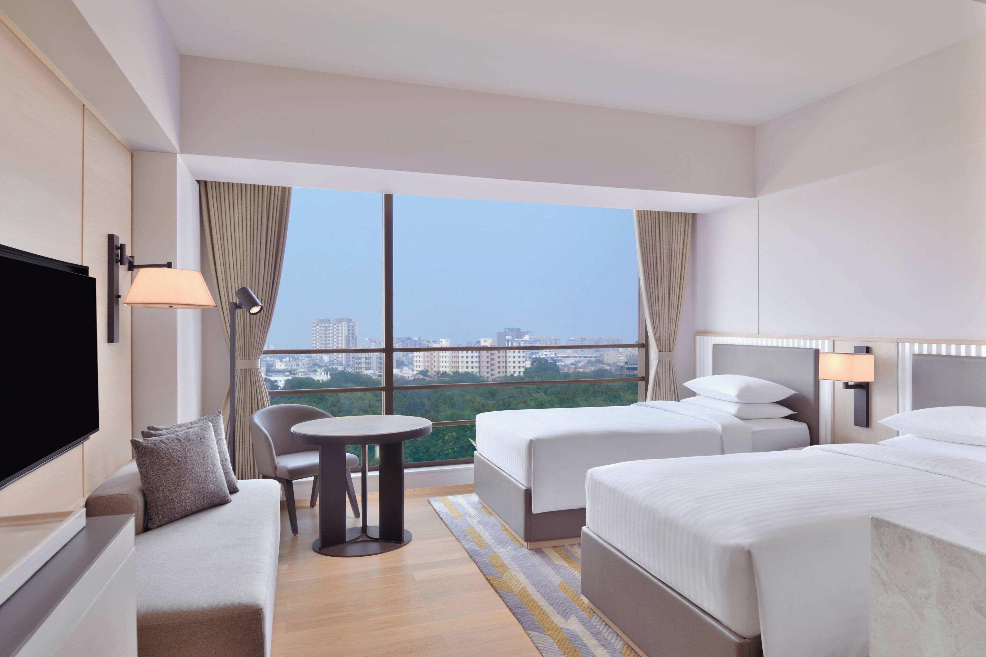 A hotel room with two beds, a sofa, a TV, a small table, and a city view through large windows.