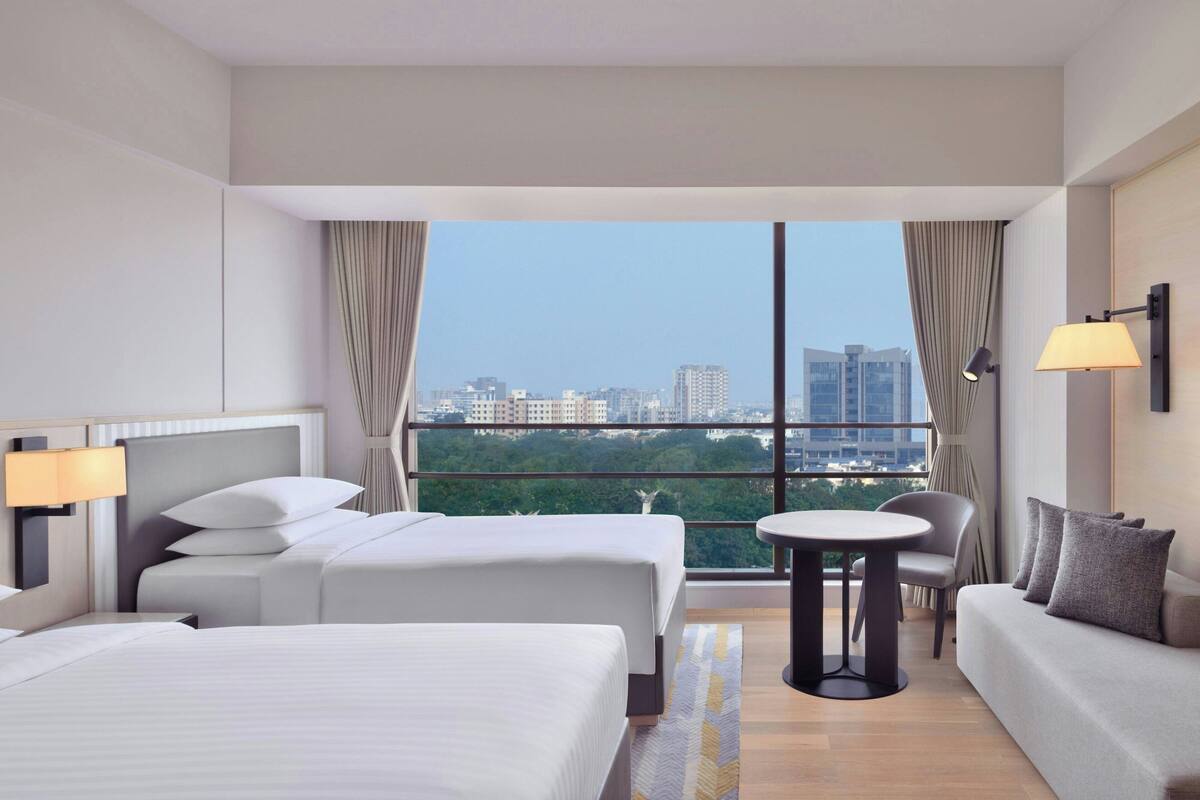 Ein Hotelzimmer mit zwei Betten, einem Schlafsofa, einem kleinen Tisch und Blick auf eine Stadtlandschaft.