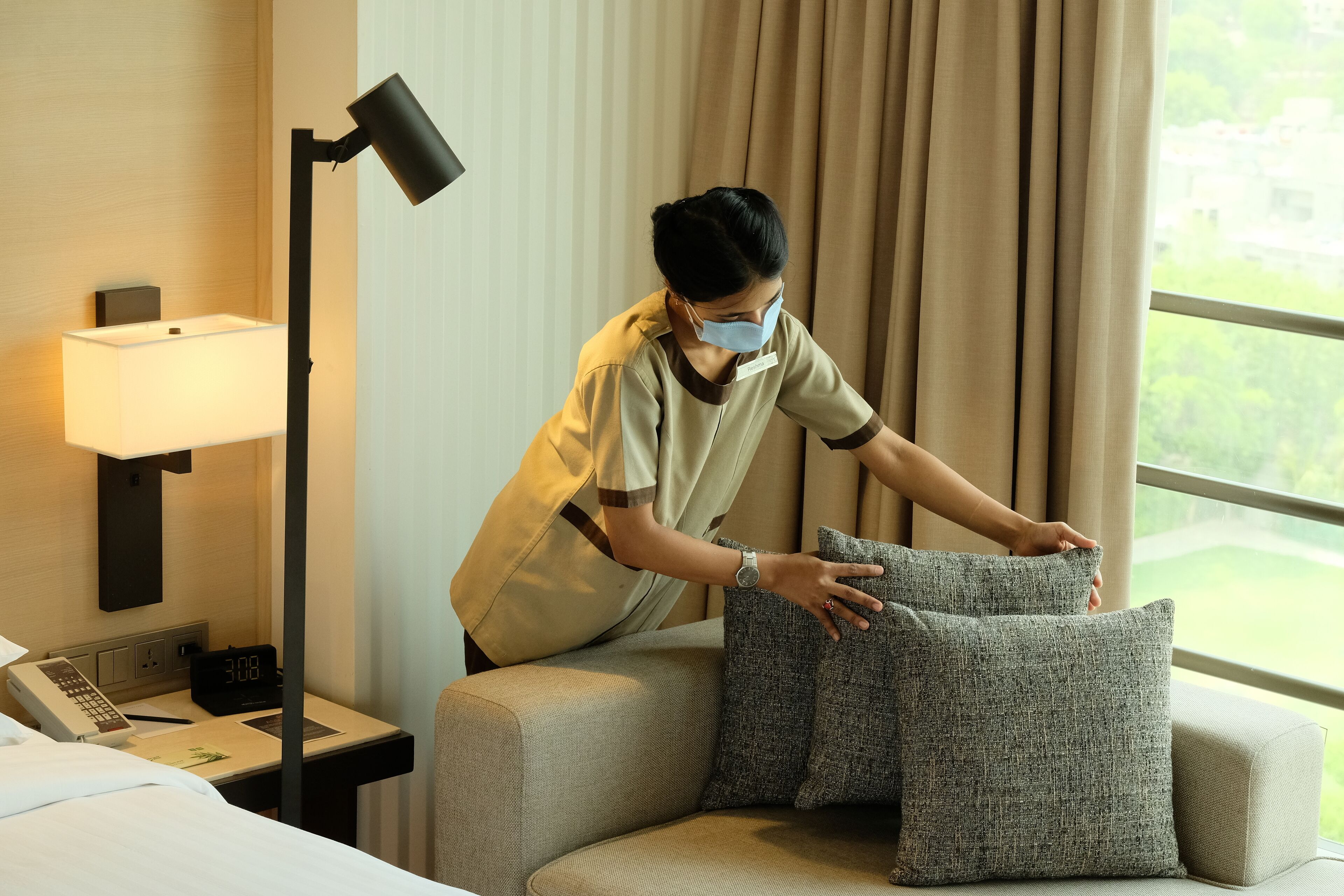 Habitación de hotel con cama, mesita de noche con lámpara, reloj digital y vistas al exterior con vegetación.