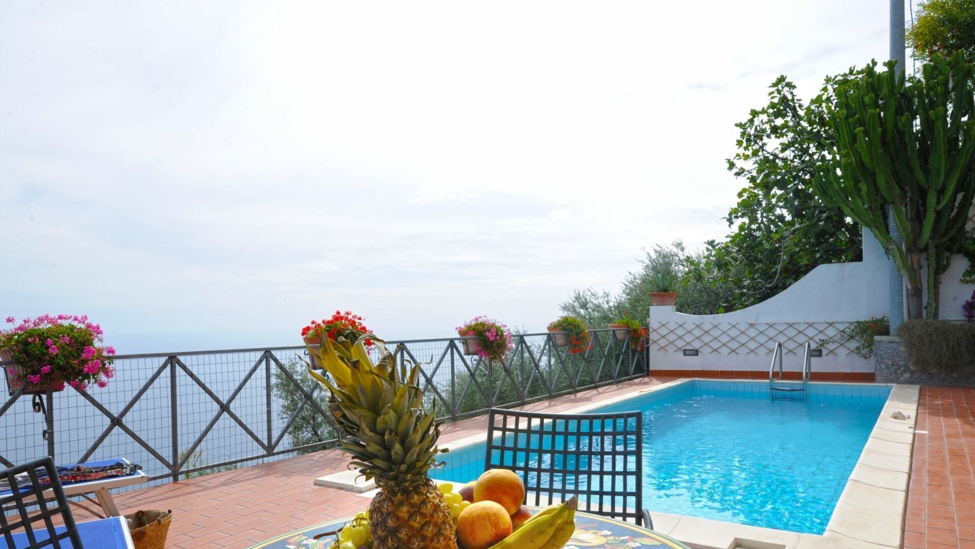 A poolside area with a view of the sea, a table with fruits, and hanging flower baskets.
