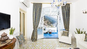 A living room with a TV, a white armchair, a sofa, a chandelier, and a view of a balcony with a pool.