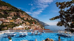 A pool area with lounge chairs and tables, overlooking a coastal town with buildings on a hillside.