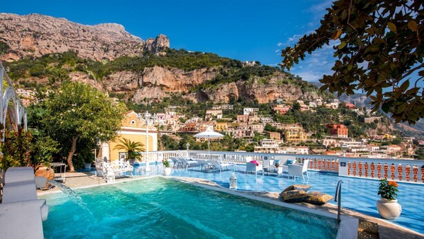 A pool area with clear blue water, white lounge chairs, and a view of a hillside town with buildings and greenery.
